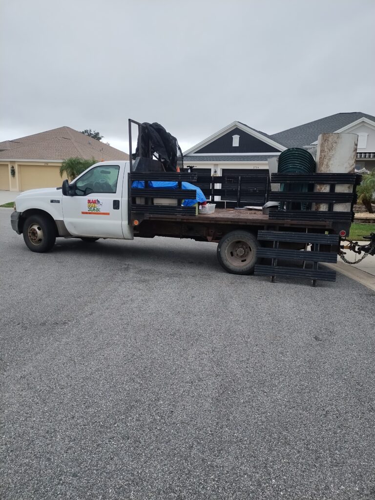 A photograph of a white Ford F-Series flatbed work truck for 'Slam Dunk Junk Removal', parked on a suburban street. The company logo, which includes a basketball and hoop, is visible on the cab door. The black-slat flatbed is heavily loaded with various items, including a stack of green plastic patio chairs, an aged metal tank, and blue and black tarps. The background features residential houses under an overcast sky.
